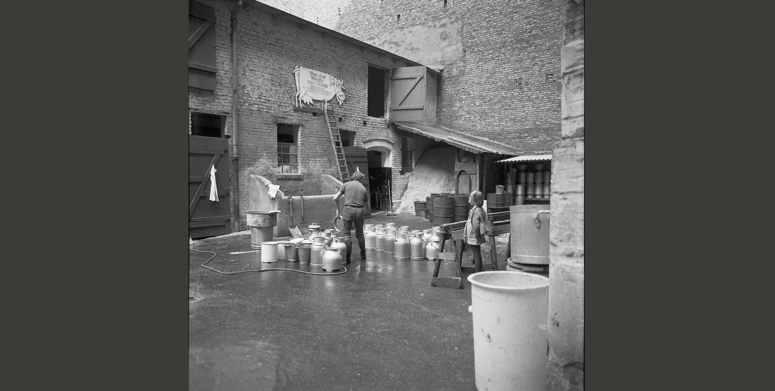 Historisches Schwarz-Weiß-Foto: ein Mann in einem Innenhof, in dem diverse Milchkannen stehen. An einer Remise im Hintergrund ist ein großes Schild in Form einer Kuh zu sehen. Aufschrift: Von der Weide zur Milch." Foto: Landesarchiv Berlin