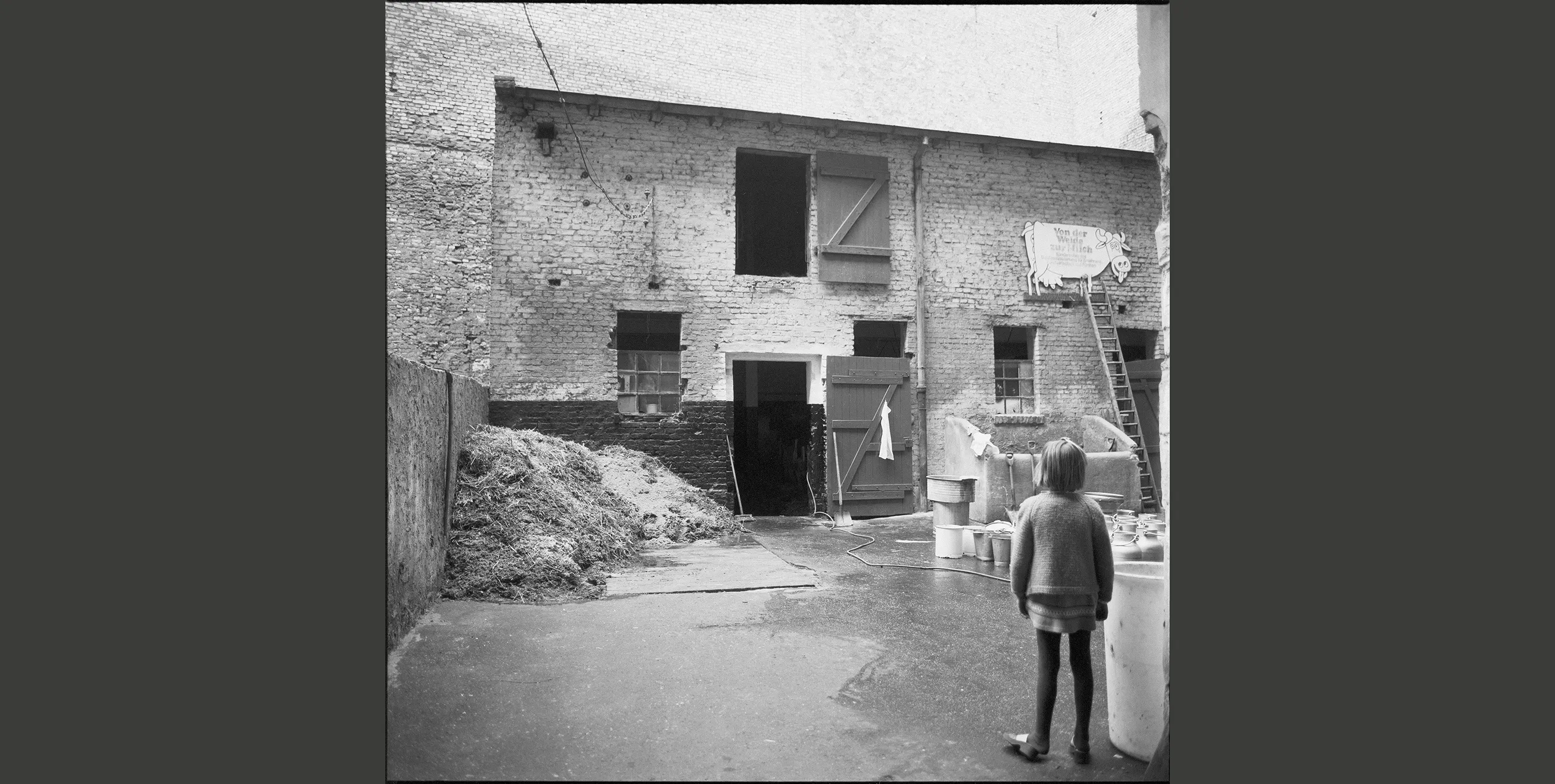 Historisches Schwarz-Weiß-Foto: Ein Kind steht in einem Innenhof, im Hintergrund ist ein landwirtschaftlicher Betrieb zu sehen. In einer Hecke liegt ein größerer Haufen Stroh. Foto: Landesarchiv Berlin