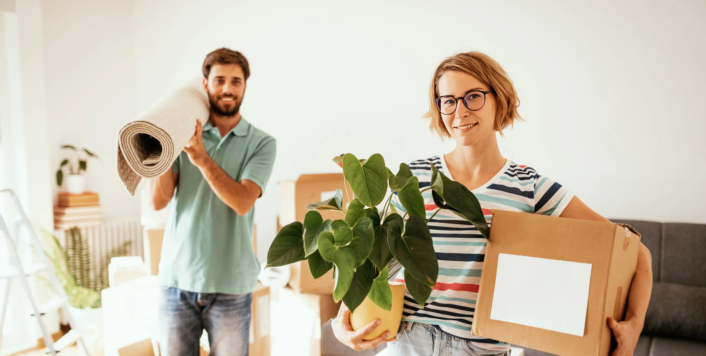Paar steht in neuer Wohnung mit Teppich auf der Schulter und Pflanze und Umzugskiste auf dem Arm.