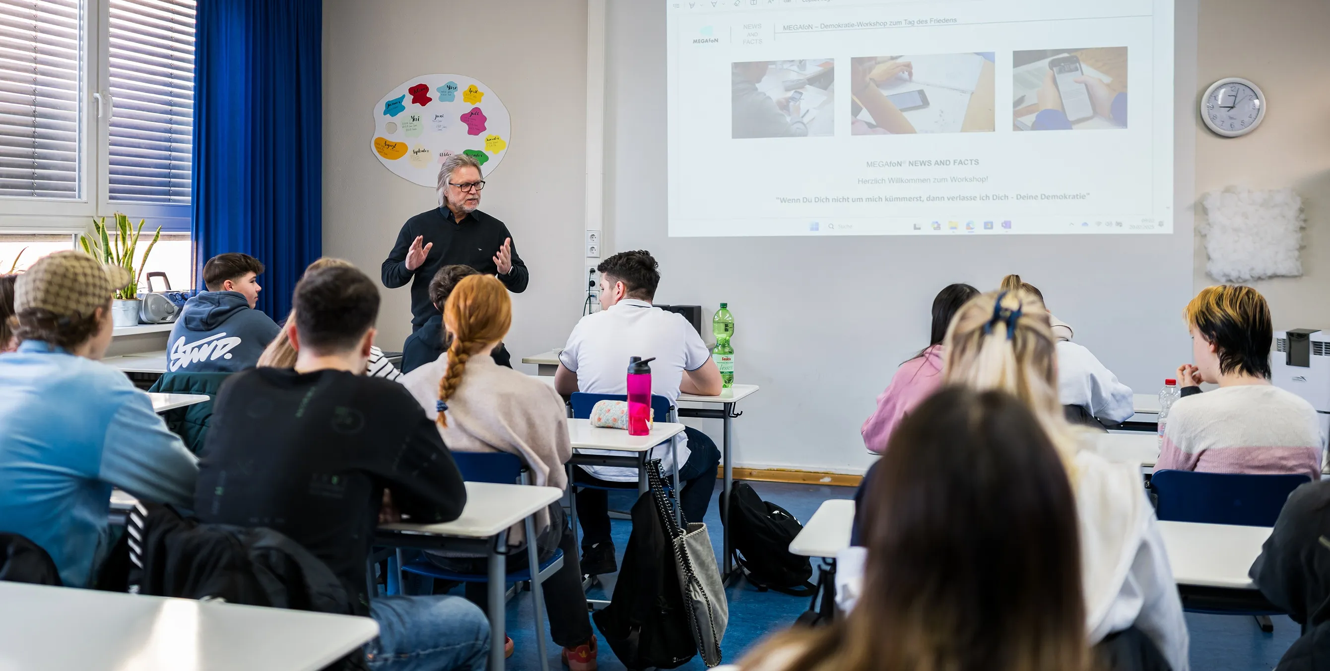 Aufnahme von hinten: Mehrere SchülerInnen sitzen in einer Schulklasse an ihren Tischen und schauen nach vorn. Dort hält ein Workshopleiter einen präsentationsgestützten Vortrag. Foto: City-Press GmbH