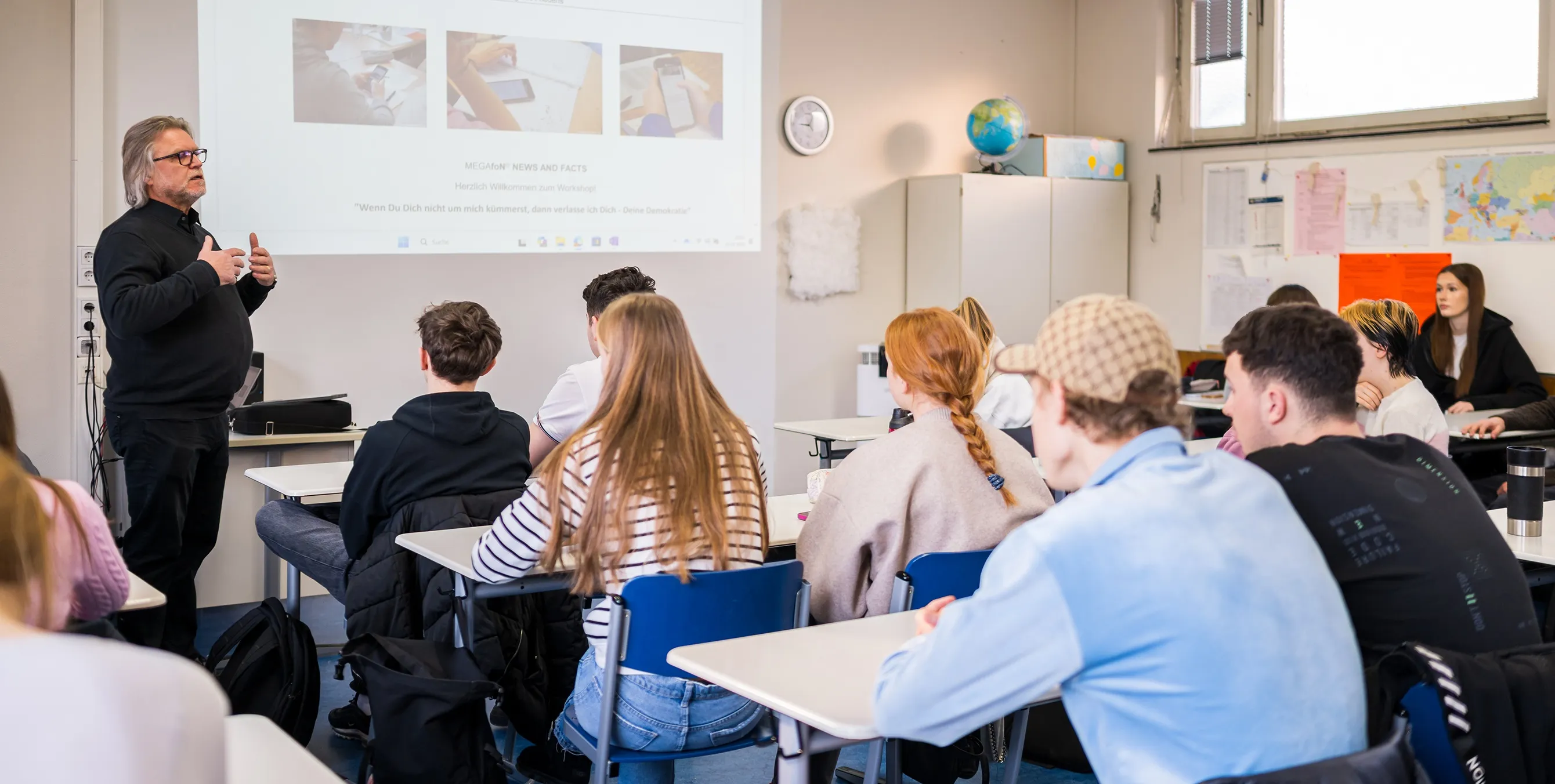 Aufnahme von hinten: Mehrere SchülerInnen sitzen in einer Schulklasse an ihren Tischen und schauen nach vorn. Dort hält ein Workshopleiter einen präsentationsgestützten Vortrag. Foto: City-Press GmbH