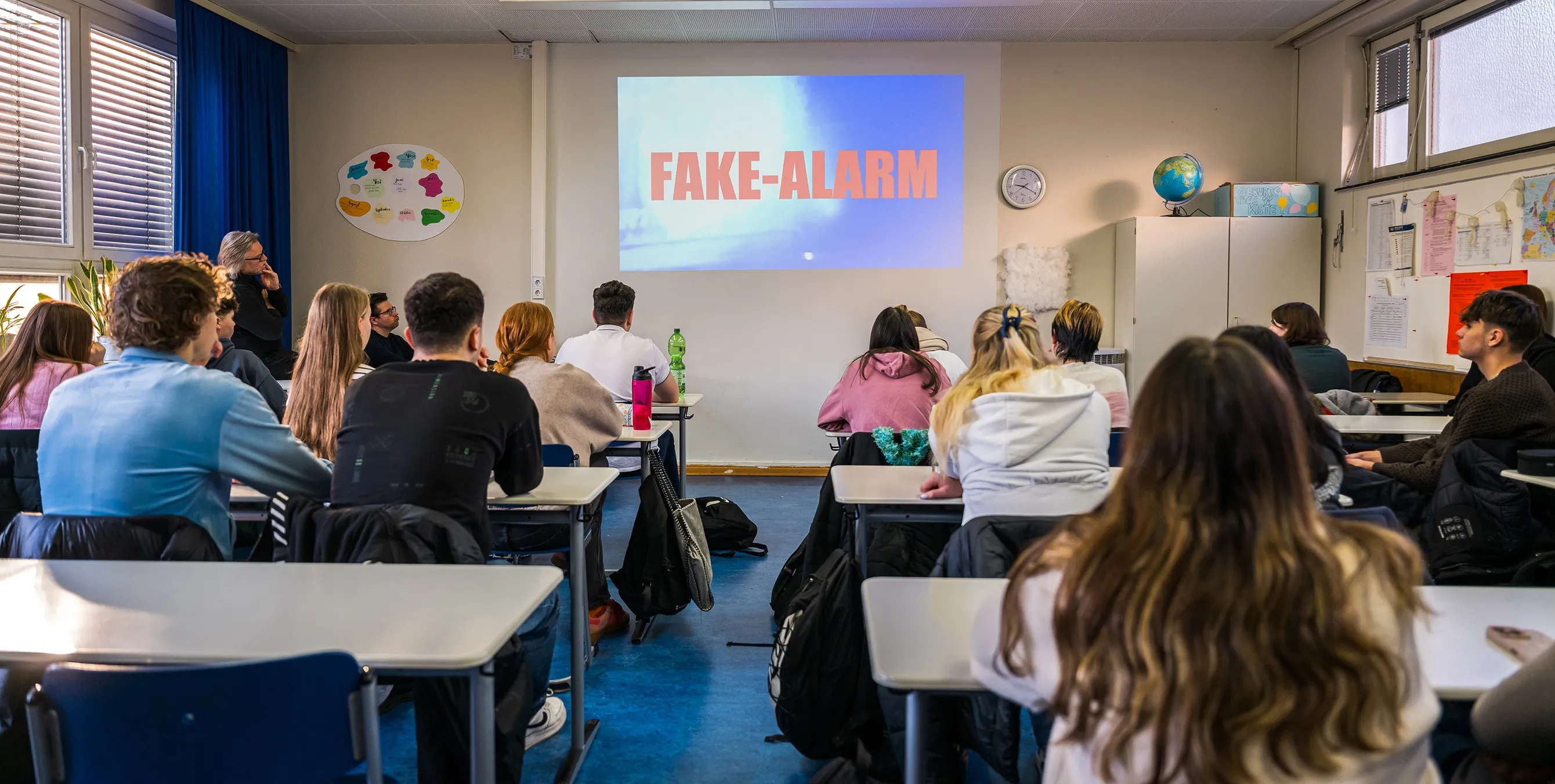 Aufnahme von hinten: Mehrere SchülerInnen sitzen in einer Schulklasse an ihren Tischen und schauen nach vorn. Dort ist in großen, roten Buchstaben der Begriff "Fake-Alarm" an die Wand projiziert. Foto: City-Press GmbH