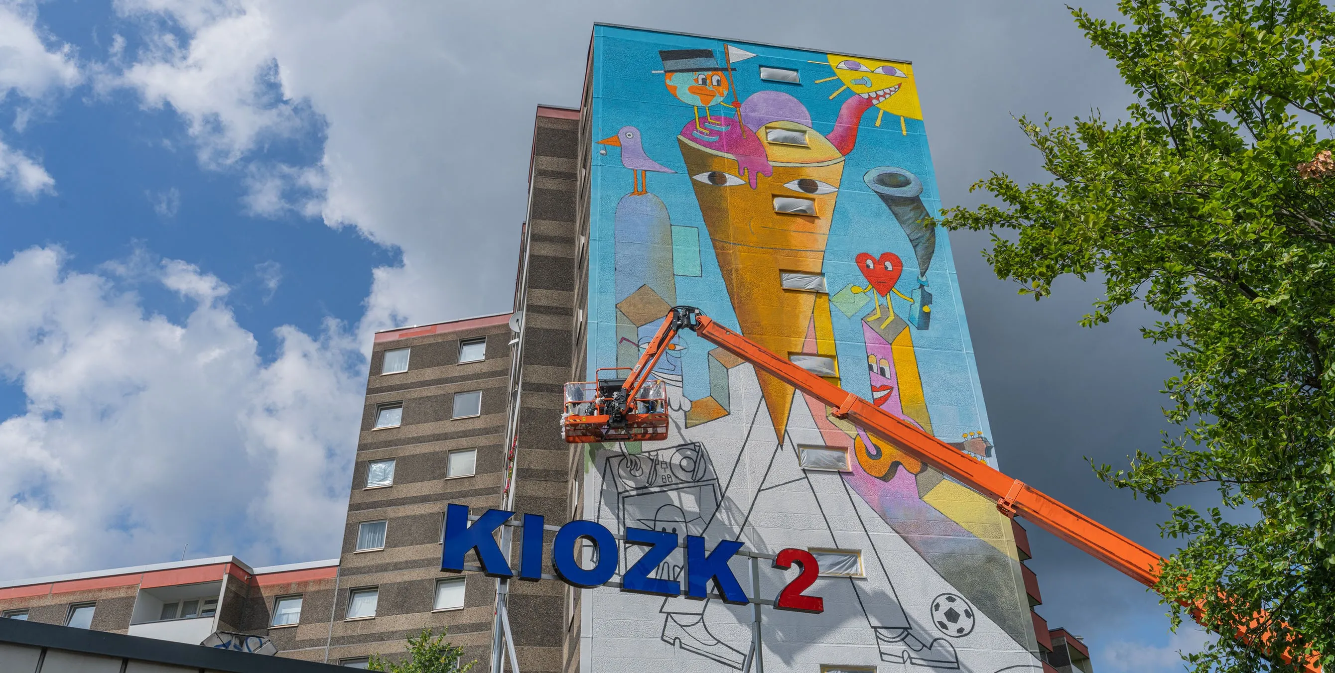 Ansicht von unten: Die Fassade eines Hochhauses während einer Bemalungsaktion. Vor dem Gebäude steht ein Kran, der die Arbeitsplattform der Künstler in die Höhe hievt. Foto: Felix Seyfert