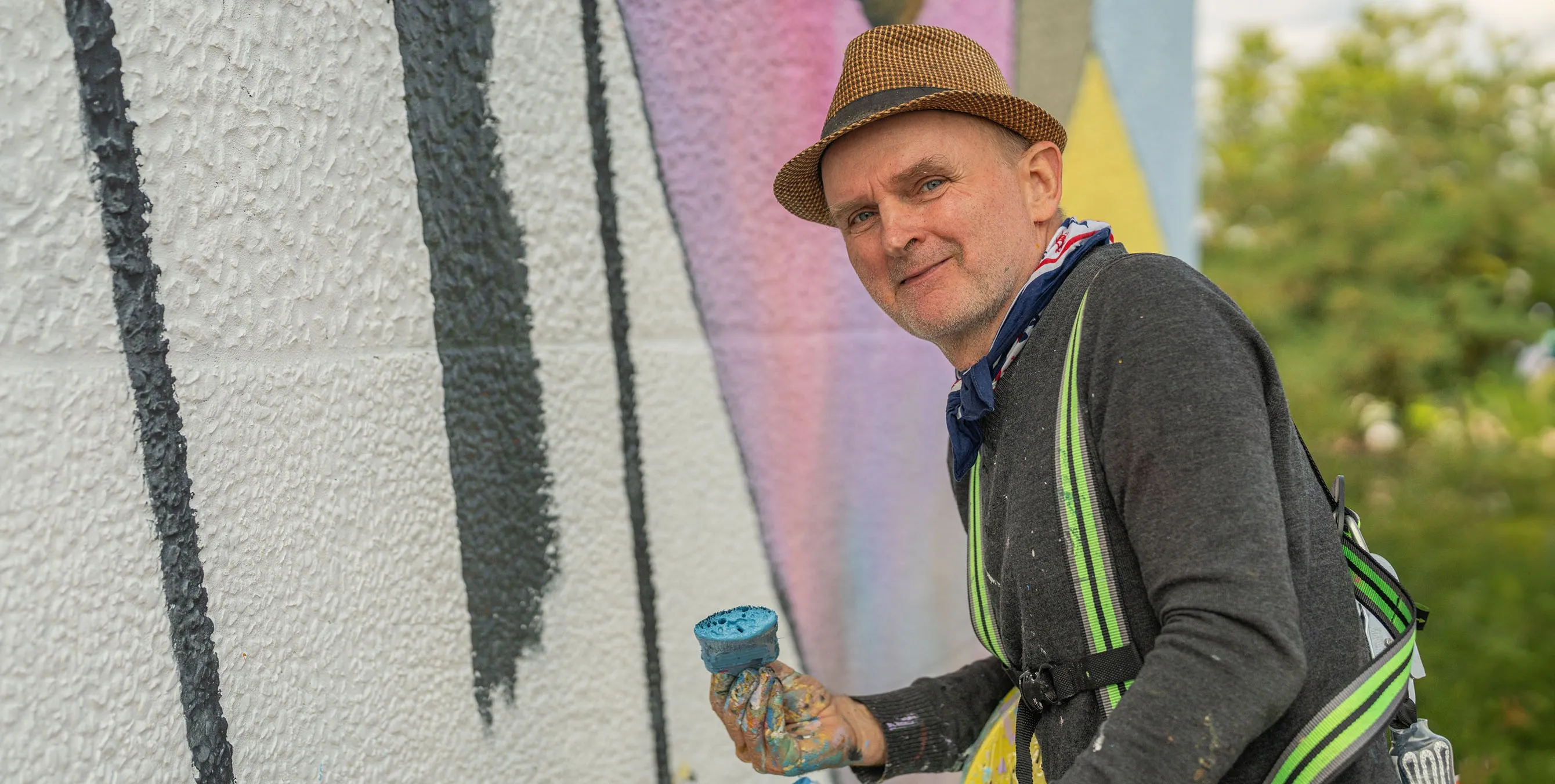 Ein Künstler mit Hut arbeitet an einer Hausfassade und schaut in die Kamera. Foto: Felix Seyfert