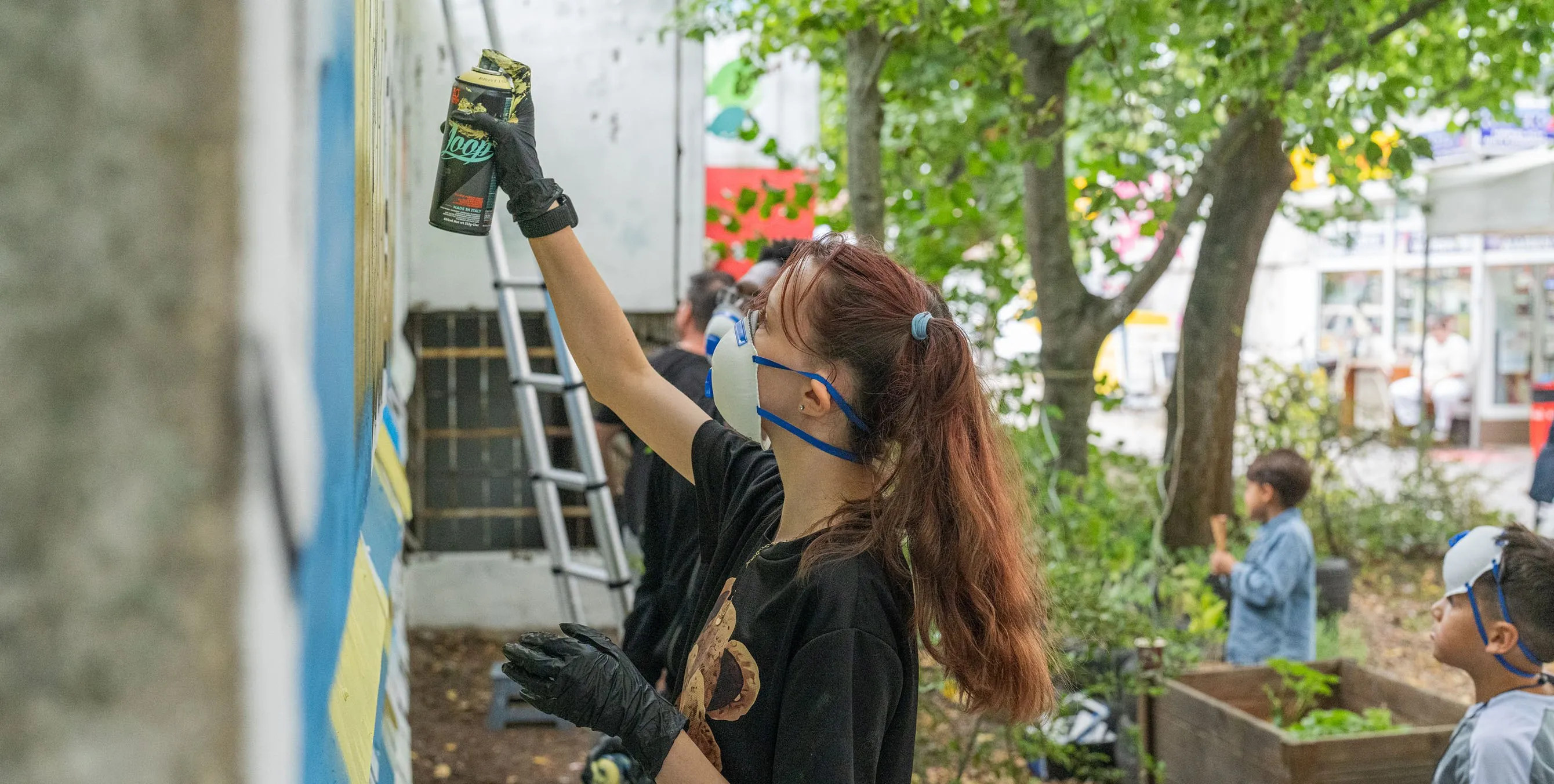 Szene im Freien: Ein junges Mädchen mit Mundschutz und Handschuhe sprüht bei einer Graffiti-Aktion etwas mit einer Spraydose an eine Wand. Foto: Felix Seyfert