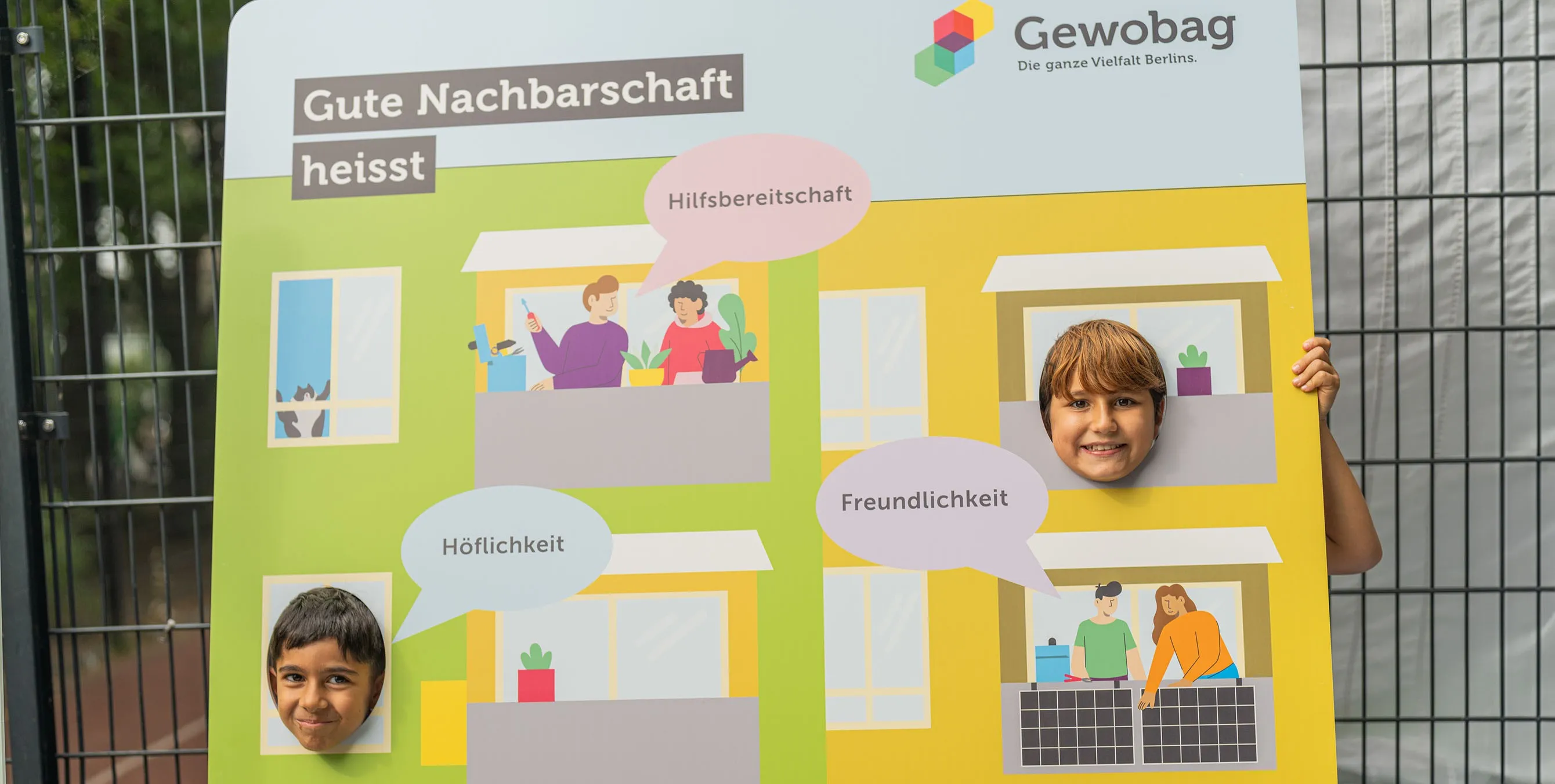 Zwei Kinder stecken ihre Köpfe durch die Löcher in einer Fotowand. Jene ist in Form einer Häuserfassade gestaltet. Darauf zu lesen ist "Gute Nachbarschaft heißt" sowie die Stichworte "Hilfsbereitschaft", "Höflichkeit" und "Freundlichkeit". Foto: Felix Seyfert