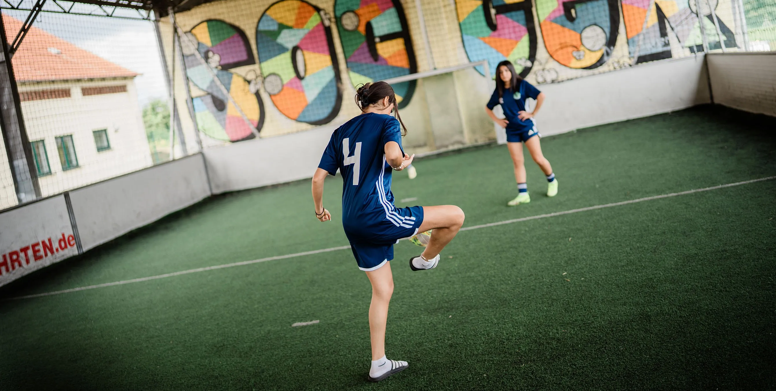 Zwei Teenagerinnen in Fußballoutfit stehen in einem überdachten Fußballkäfig und kicken. Foto: Johannes Karnauchow/3viertel3