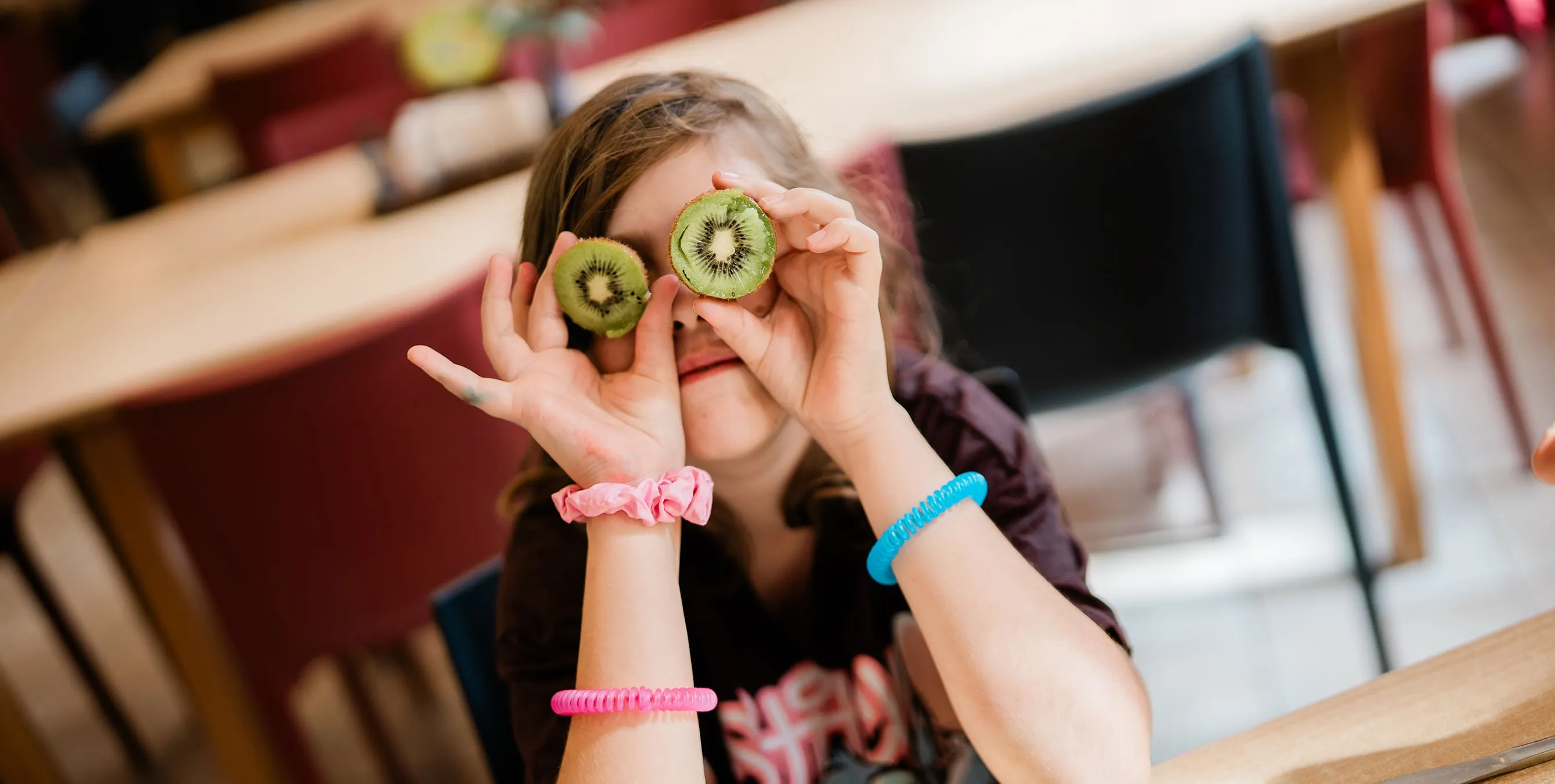 Szene aus einem Speisesaal: Ein Kind hält die beiden aufgeschnittenen Hälften eine Kiwi vor seine Augen und blickt Richtung Kamera. Foto: Johannes Karnauchow/3viertel3