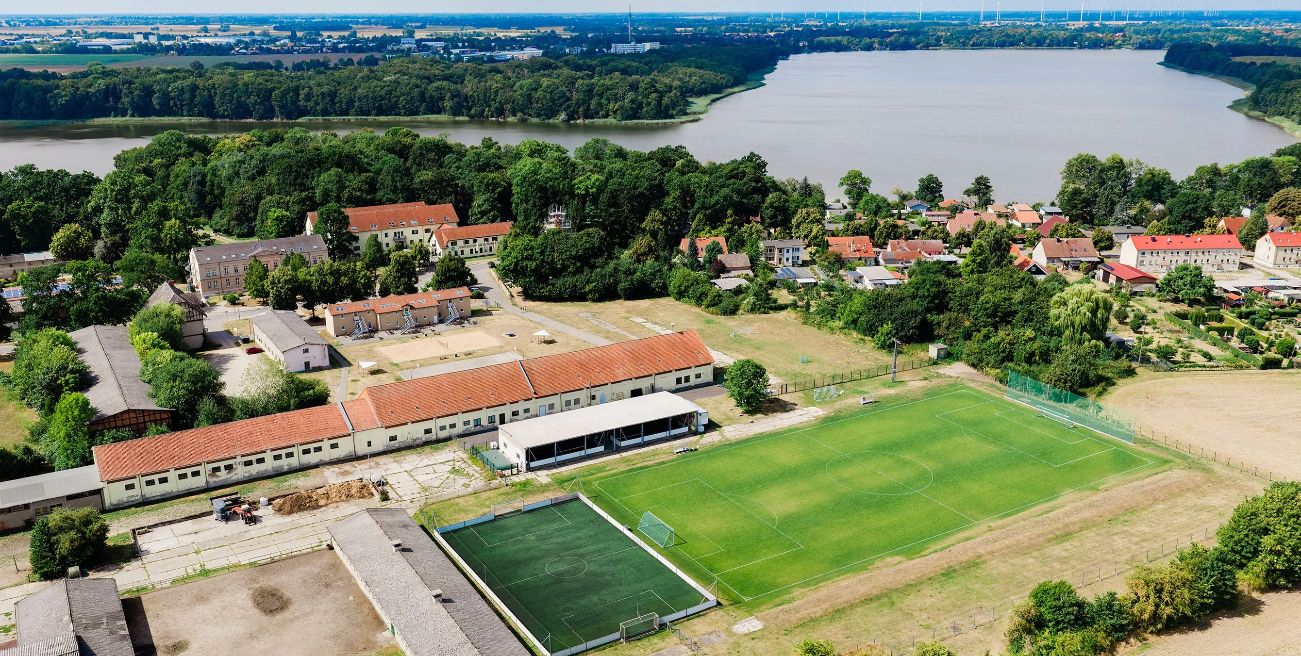 Das Jugenddorf Ruppiner See aus der Luft. Im Vordergrund sind Fußballplätze zu sehen, im Hintergrund der große Ruppiner See. Foto: Johannes Karnauchow/3viertel3