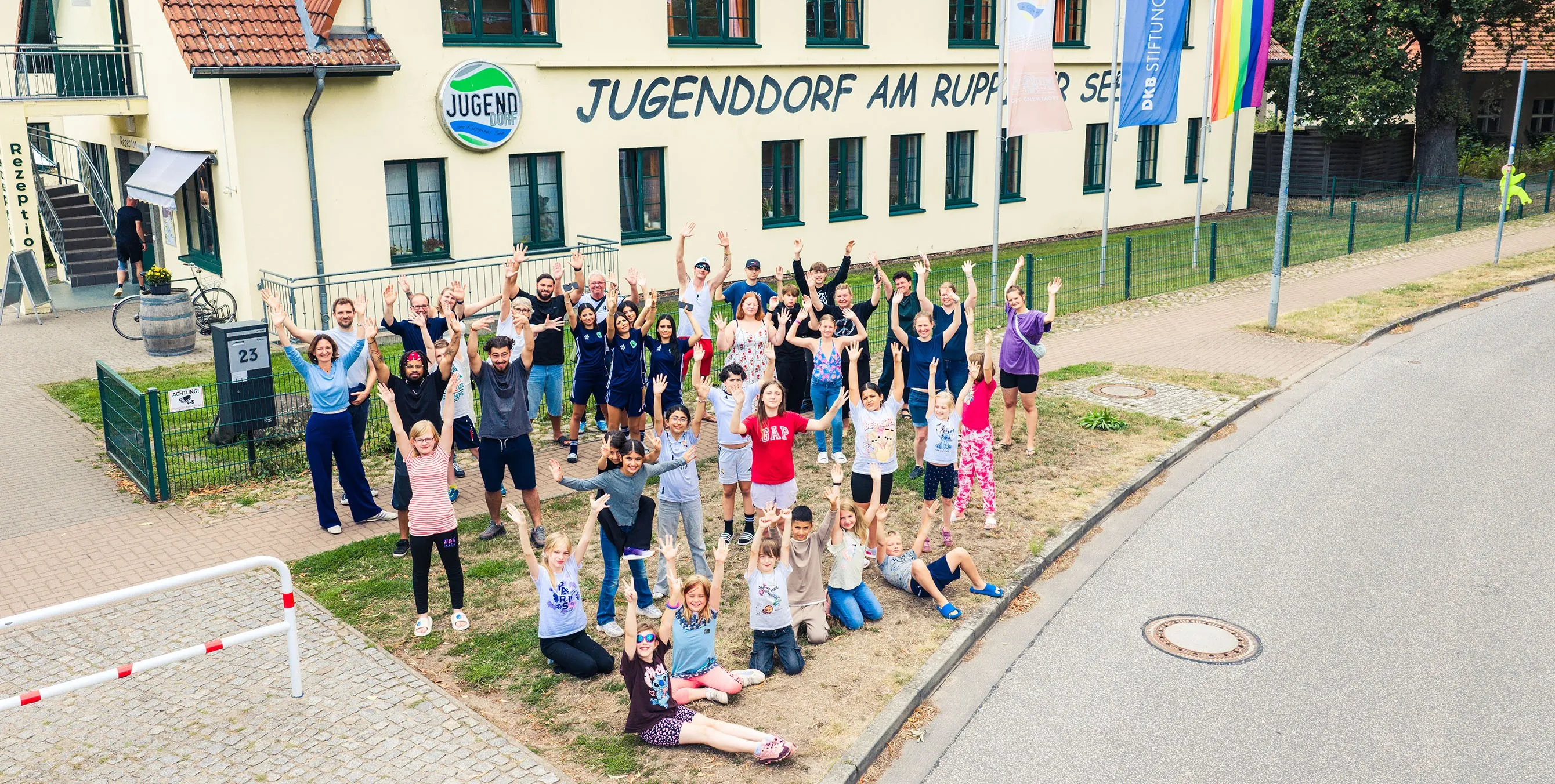 Rund 40 Personen - Kinder, Jugendliche und junge Erwachsene - stehen vor einem Gebäude mit der Aufschrift "Jugenddorf am Ruppiner See", recken die Hände in die Luft und schauen in Richtung Kameradrohne. Foto: Johannes Karnauchow/3viertel3