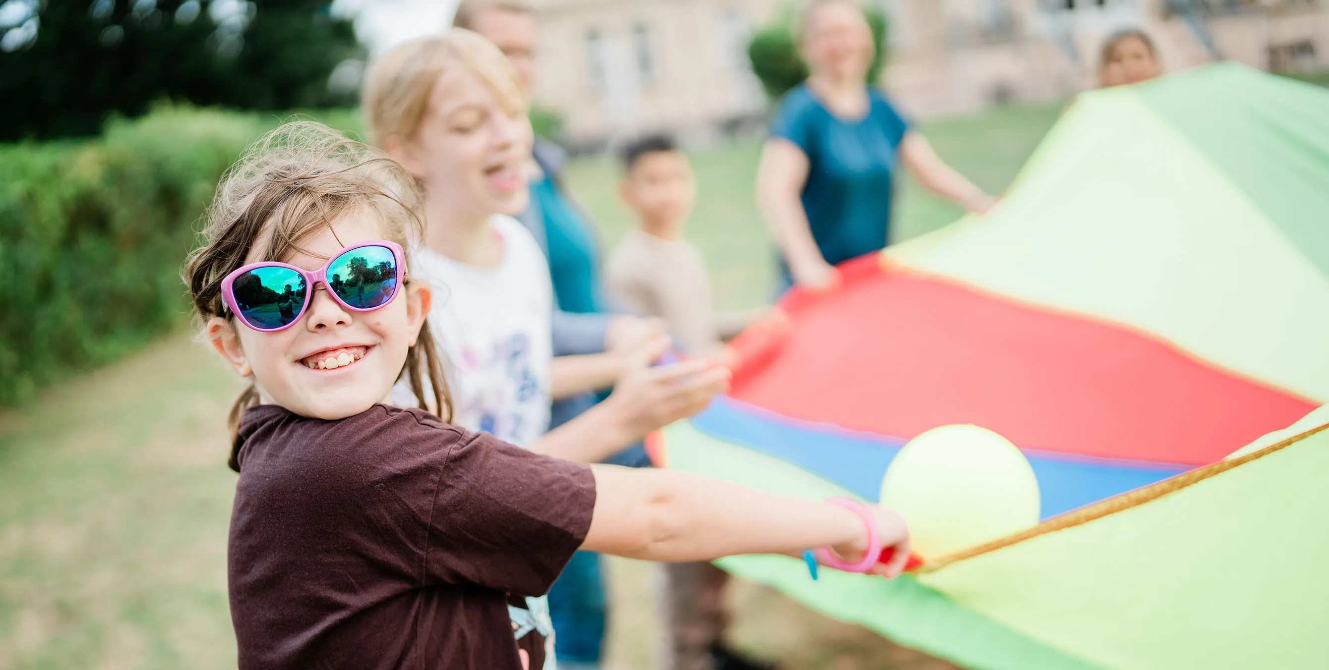 Kinder spielen im Freien mit einem bunten Schwungtuch. Ein Mädchen mit großer Sonnenbrille lächelt in die Kamera. Foto: Johannes Karnauchow/3viertel3