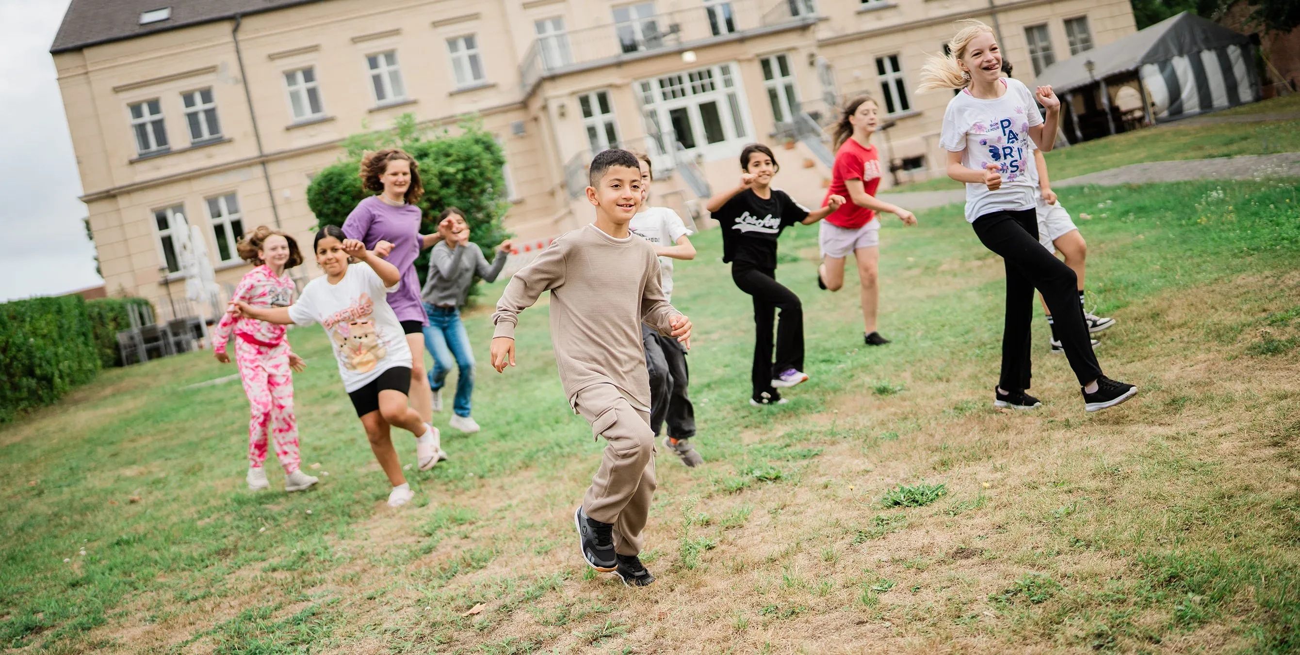 Kinder rennen im Freien lachend über eine Rasenfläche. Im Hintergrund ist ein altes Gutsgebäude zu sehen. Foto: Johannes Karnauchow/3viertel3