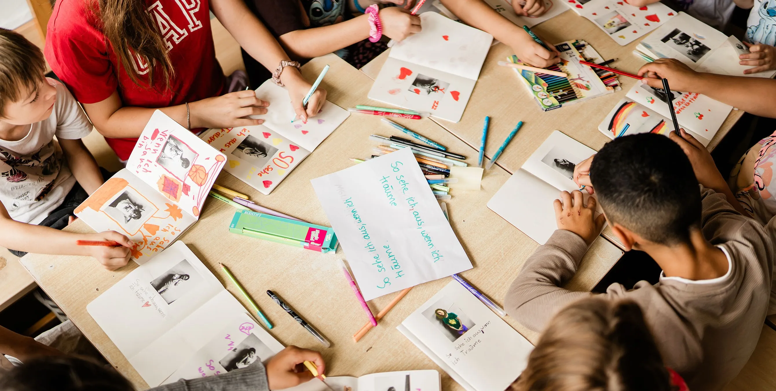 Kinder sitzen an einem Tisch und schreiben mit bunten Stiften etwas in die Hefte, die vor ihnen liegen. In der Mitte des Tisches liegt ein Zettel mit den Worten: "So sehe ich aus, wenn ich träume." Foto: Johannes Karnauchow/3viertel3