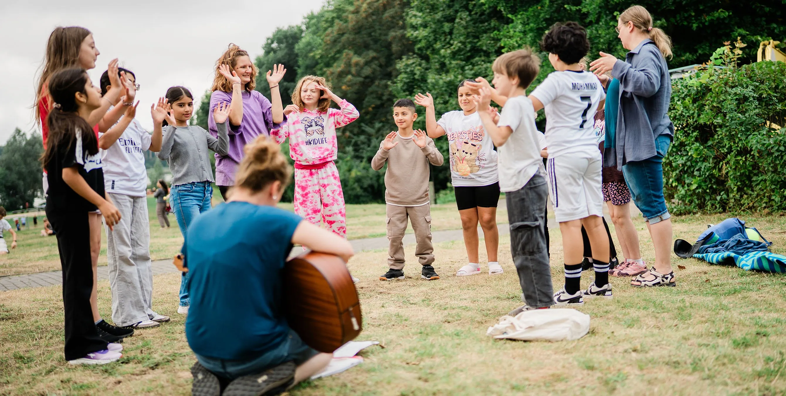 Ein Dutzend Kinder stehen im Freien im Kreis und machen Gesten mit ihren Armen. Im Vordergrund kniet eine Frau und spielt auf einer Gitarre. Foto: Johannes Karnauchow/3viertel3