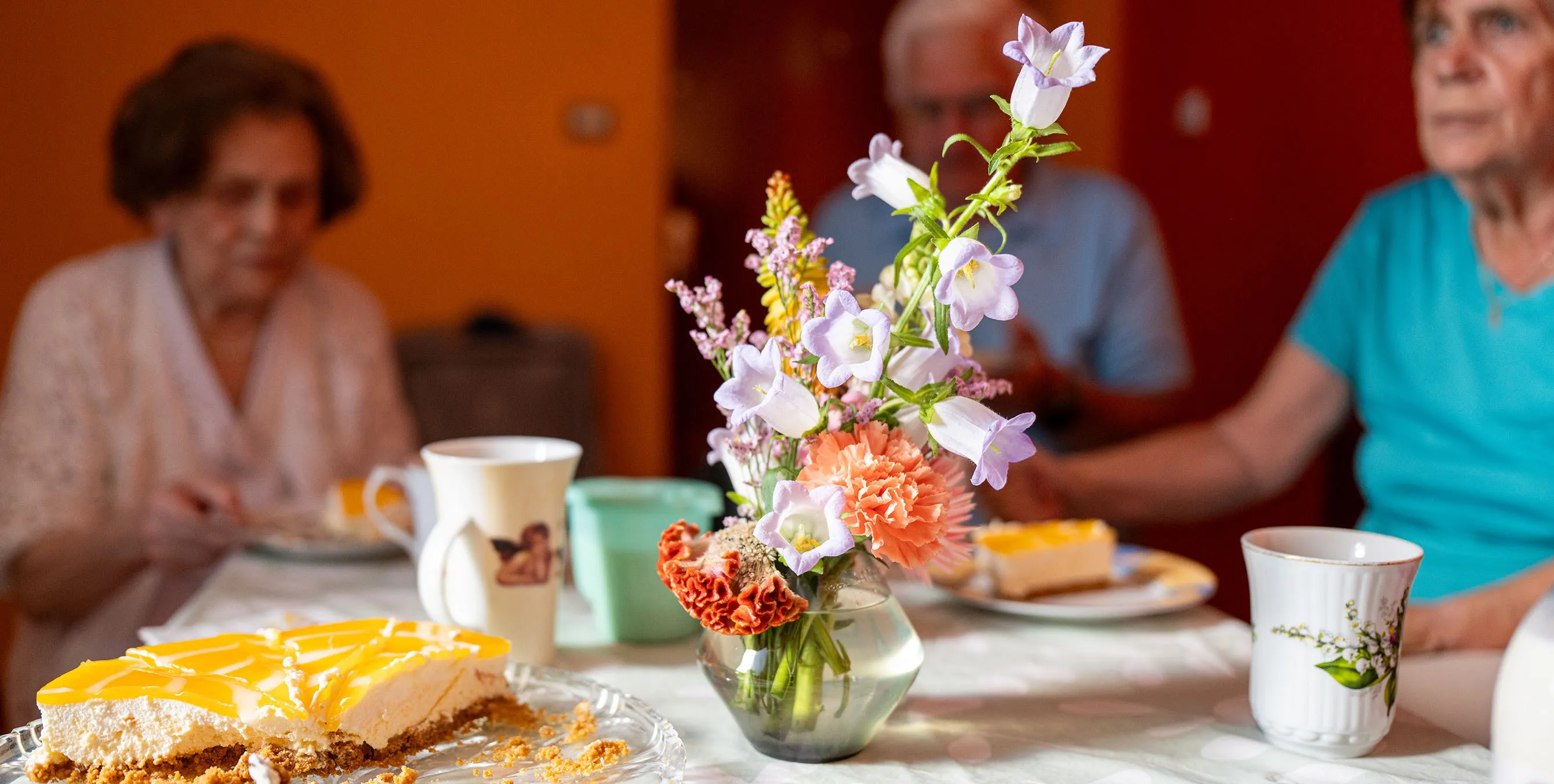 Großaufnahme: ein Blumenstrauß in einer gläsernen Vase, die auf einem gedeckten Kaffeetisch steht. Im Hintergrund sind sitzende Personen zu sehen. Foto: Ralph Maak