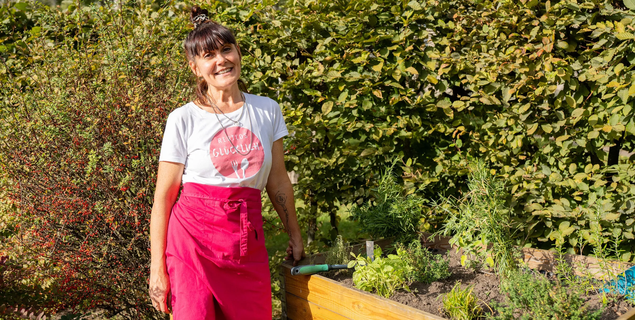 Frau mit pinker Schürze steht lächelnd am bepflanzten Hochbeet im Garten. Foto: Ralph Maak