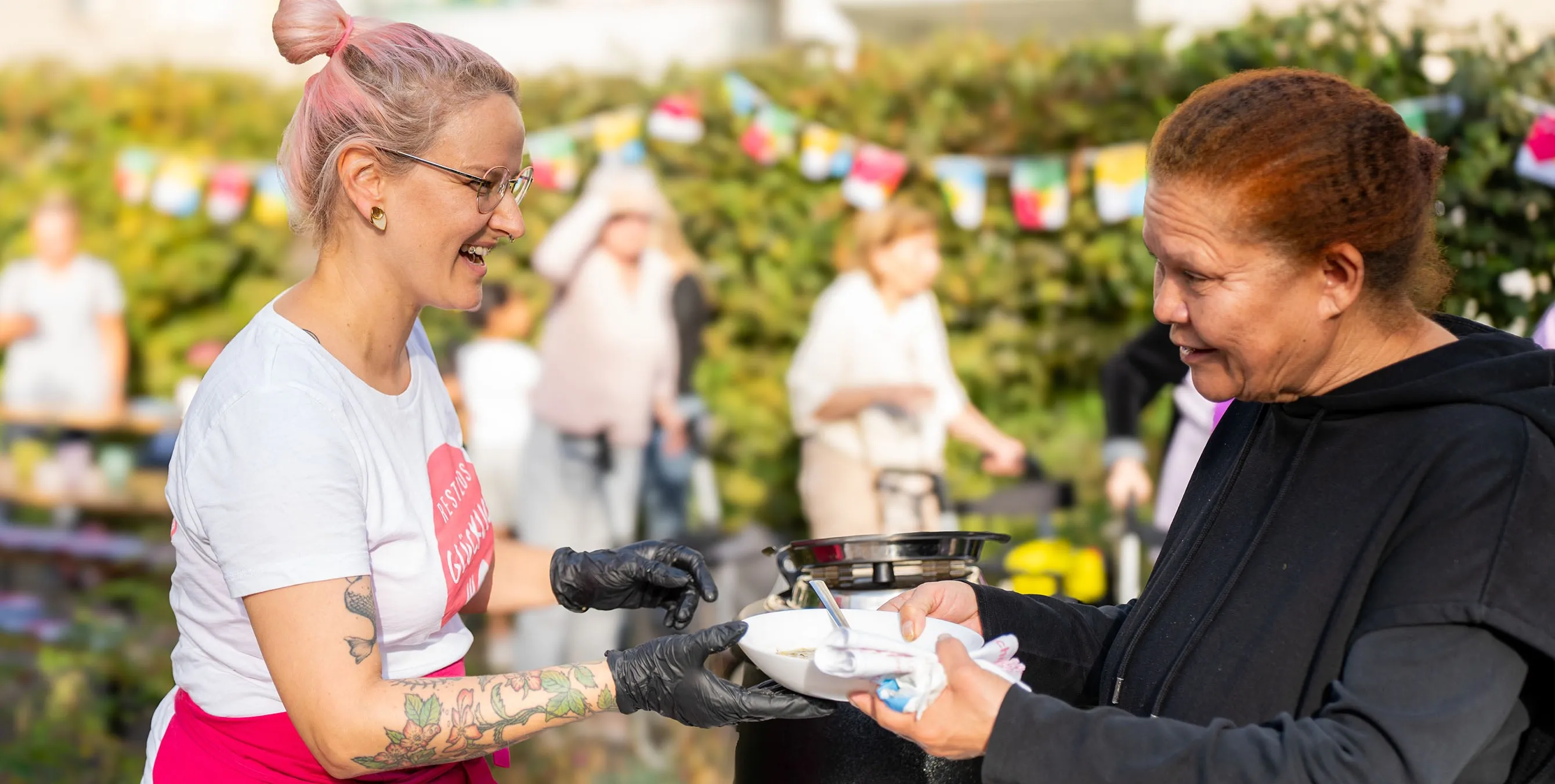Frau mit pinken Haaren serviert eine Schale Essen an eine Besucherin im Freien. Foto: Ralph Maak