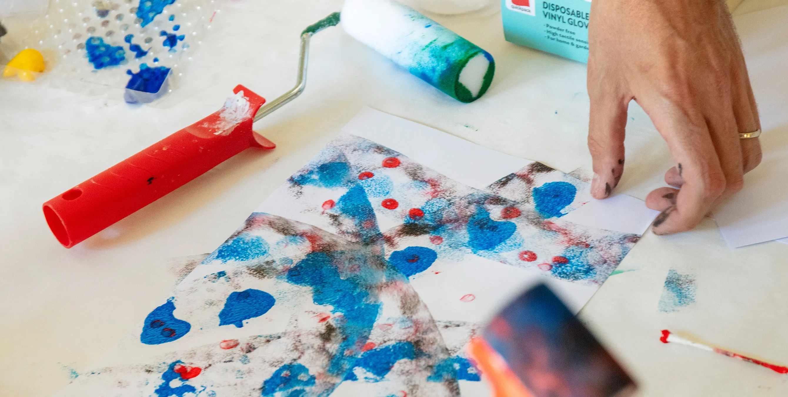 Nahaufnahme: Ein Hand greift auf einem Maltisch zu einem bemalten Blatt Papier. Daneben sind Arbeitsutensilien wie Farbwanne und Farbrollen zu sehen. Foto: Ludger Paffrath