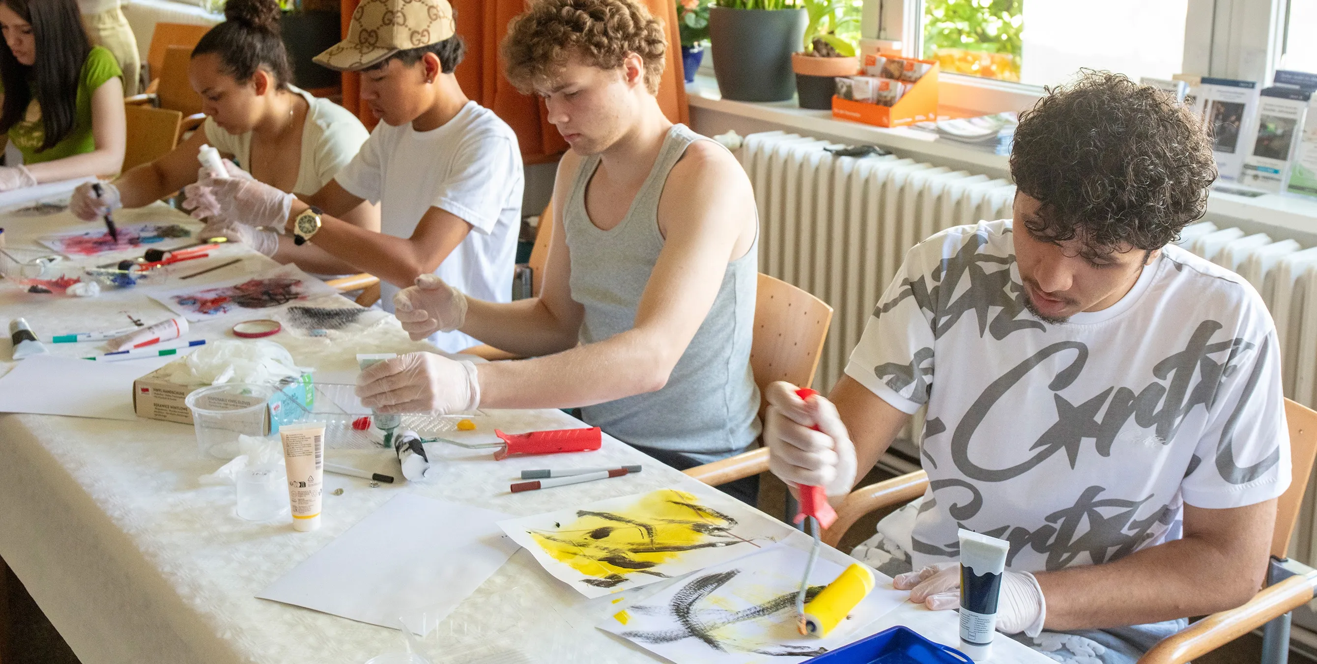 Mehrere Jugendliche sitzen an einer langen Tafel und malen mit verschiedenen Farben und Materialien und Arbeitsutensilien. Foto: Ludger Paffrath