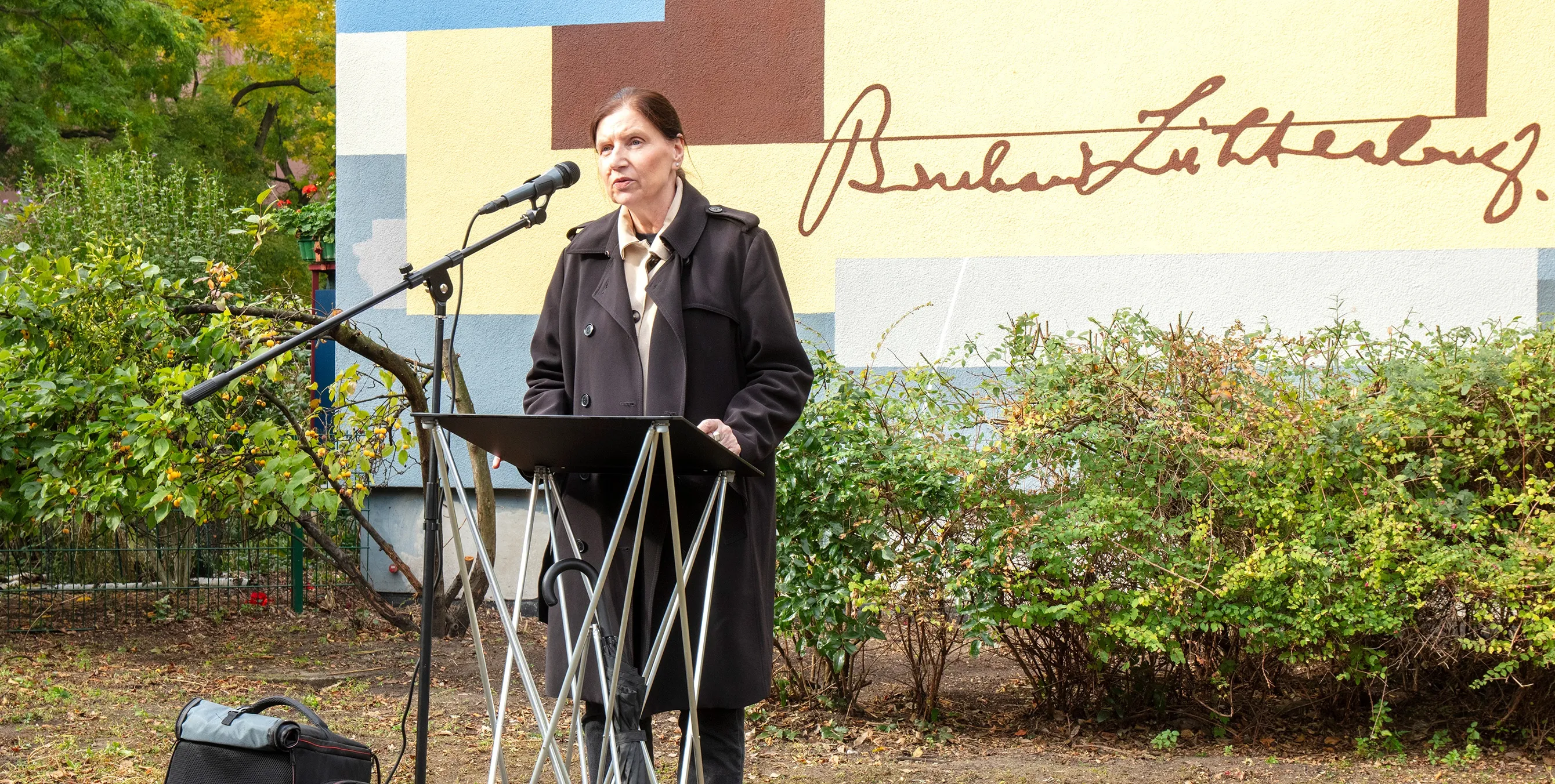 Eine Frau mit schwarzem Mantel steht an einem Rednerpult mit Mikrofon im Freien und hält eine Rede. Im Hintergrund ist eine kunstvoll gestaltete Häuserwand zu sehen. Foto: Ludger Paffrath