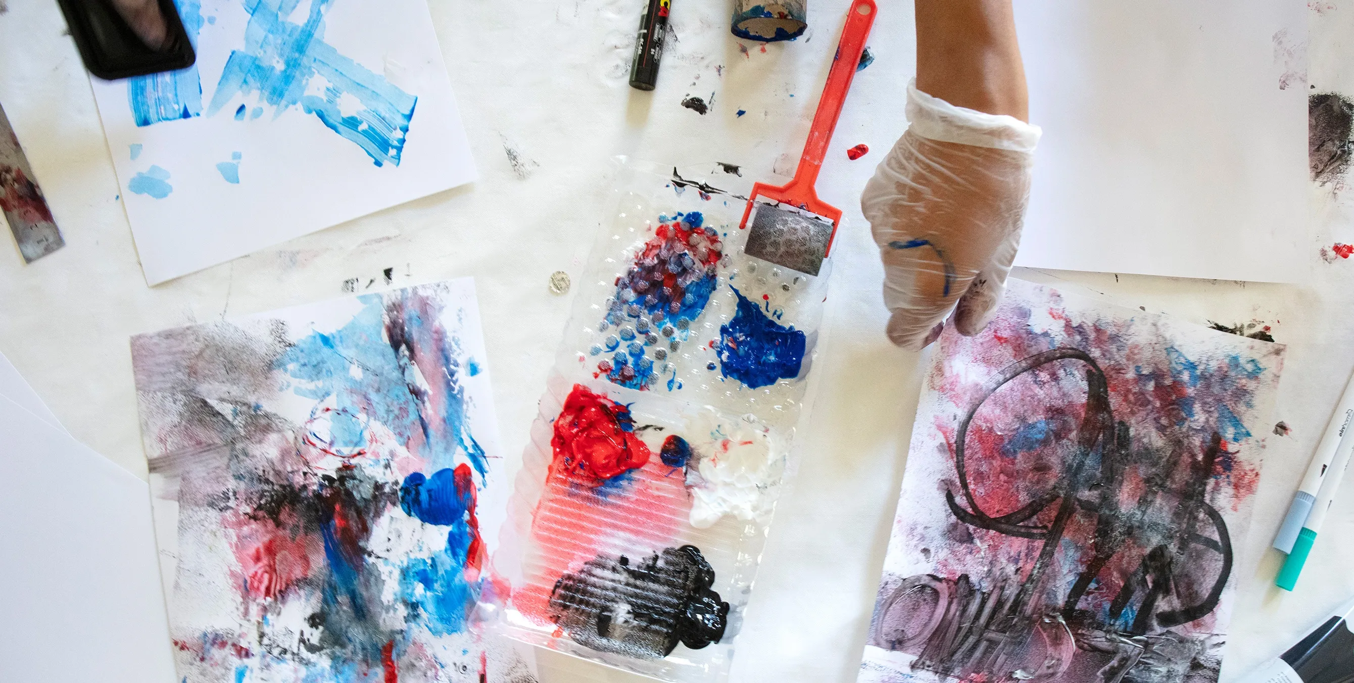 Nahaufnahme: Ein Hand greift auf einem Maltisch zu einem bemalten Blatt Papier. Daneben sind Arbeitsutensilien wie Farbwanne und Farbrollen zu sehen. Foto: Ludger Paffrath